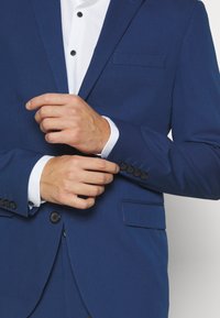 Karališkai mėlynas švarkas su notch tipo atvartais, vienu mygtuku ir šoniniu kišeniu; turi juodus mygtukus ir baltą marškinių. 