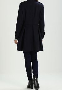 Abrigo de lana azul marino con un torso ajustado y falda acampanada. Presenta hombreras, acentos de botones y una textura suave.