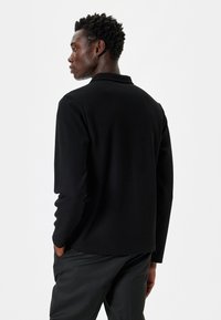 Zwarte langetermijnshirt met een gestructureerde stof en een ontspannen pasvorm. Kenmerkt zich door een kraag, subtiele stiksel details en een rechte zoom.