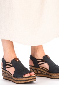 Black wedge sandals with a cork sole, featuring an open toe, crisscross straps, and a small brown accent fastener on the side.