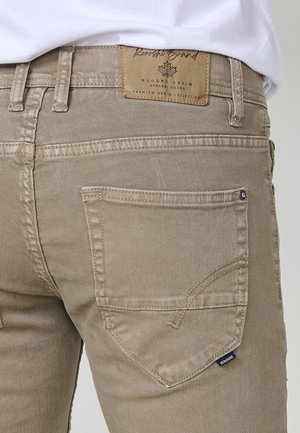 Beige Jeans aus Denim mit gestickten Details, einer Gesäßtasche und einem ledernen Etikett am Bund mit der Aufschrift "Rugged Grand". Glatte Textur.
