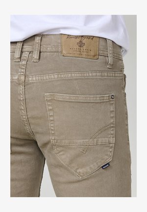 Beige denim jeans with stitched detail, a back pocket, and a leather label on the waistband marked "Rugged Grand." Smooth texture.