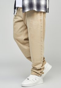 Pantaloni beige, larghi, con tasche laterali, realizzati in un tessuto resistente. Abbinati a sneakers bianche con un design testurizzato e chiusura con lacci.
