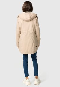 Beige parka met gevoerde capuchon, voorzien van een trekkkoord en een patch nabij de zoom. Gecombineerd met donkerblauwe jeans en witte sneakers.