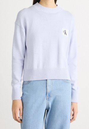 Žena nosí světle fialový žebrovaný svetr s dlouhým rukávem a malou nášivkou s logem CK na hrudi a světle modré džíny s vysokým pasem.