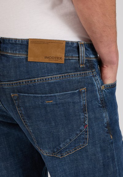 Person wearing blue denim jeans with a tan leather Incotex label on the waistband and a white shirt tucked in.