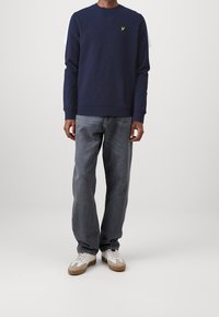 Felpa blu navy con scollo girocollo, caratterizzata da un piccolo logo giallo. Abbinata a jeans denim grigio chiaro e sneakers bianche con dettagli marroni.