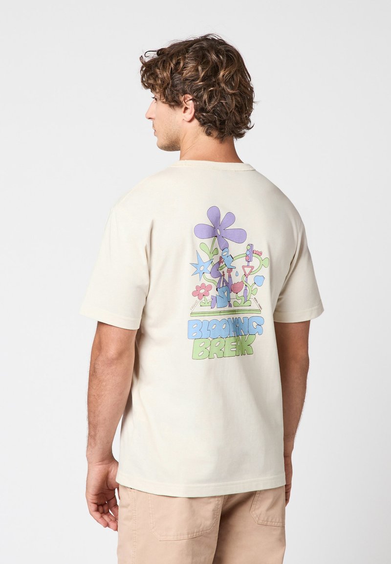 Homme portant un t-shirt beige avec un motif floral coloré et le texte « Blooming Break » au dos, vu de dos, short beige.