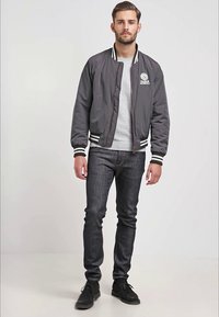 Šedá bomber bunda s bílými a černými pruhy na manžetách, nošena přes šedé tričko, v kombinaci s tmavými džíny a černými botami.
