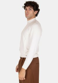 Maglione bianco a collo alto con coste in tessuto leggero, dalla silhouette aderente e maniche lunghe. Abbinato a pantaloni marroni.