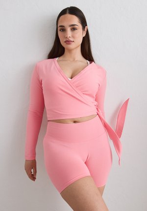 Vrouw draagt een roze wikkelstijl lange mouwen top en bijpassende hoge taille biker shorts, poserend tegen een witte muur.