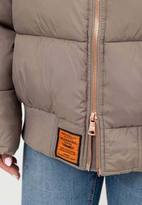 Giubbotto bomber imbottito in taupe con zip rosegold, polsini a coste e un'etichetta arancione con la scritta "BOMBERS ORIGINAL." Jeans di denim in vista.