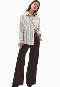 Gestreifte Hemdbluse in Schwarz und Weiß, lockere Passform, mit einer Brusttasche. Kombiniert mit hoch taillierten, weit geschnittenen braunen Hosen.