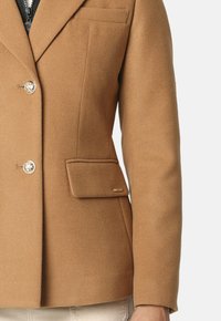 Blazer de lana marrón con cuello de muesca, dos cierres de botón dorados y un bolsillo en el pecho con un sutil acento de logo.