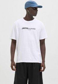 Balta kokvilnas T krekli ar "JACK&JONES" melnā un pelēkā krāsā, īsām piedurknēm, valkāta ar melnu šortu un zilu cepuri.