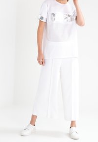 Bílý texturovaný tričko s stříbrným grafickým potiskem, spárované s bílými širokými kalhotami a bílými slip-on botami.