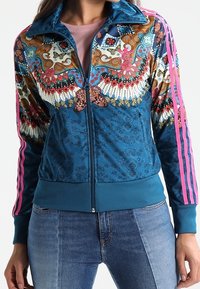 Zip-up sports jacket in blue with multicolored floral and butterfly patterns, featuring pink stripes on sleeves and a textured finish.