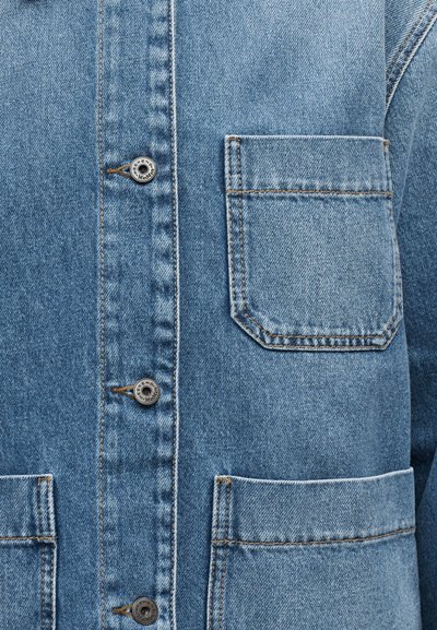 Κοντινό πλάνο μπροστινού μέρους μπλε τζιν μπουφάν με δύο κάτω τσέπες, μία τσέπη στο στήθος και μεταλλικά κουμπιά με κουμπότρυπες.