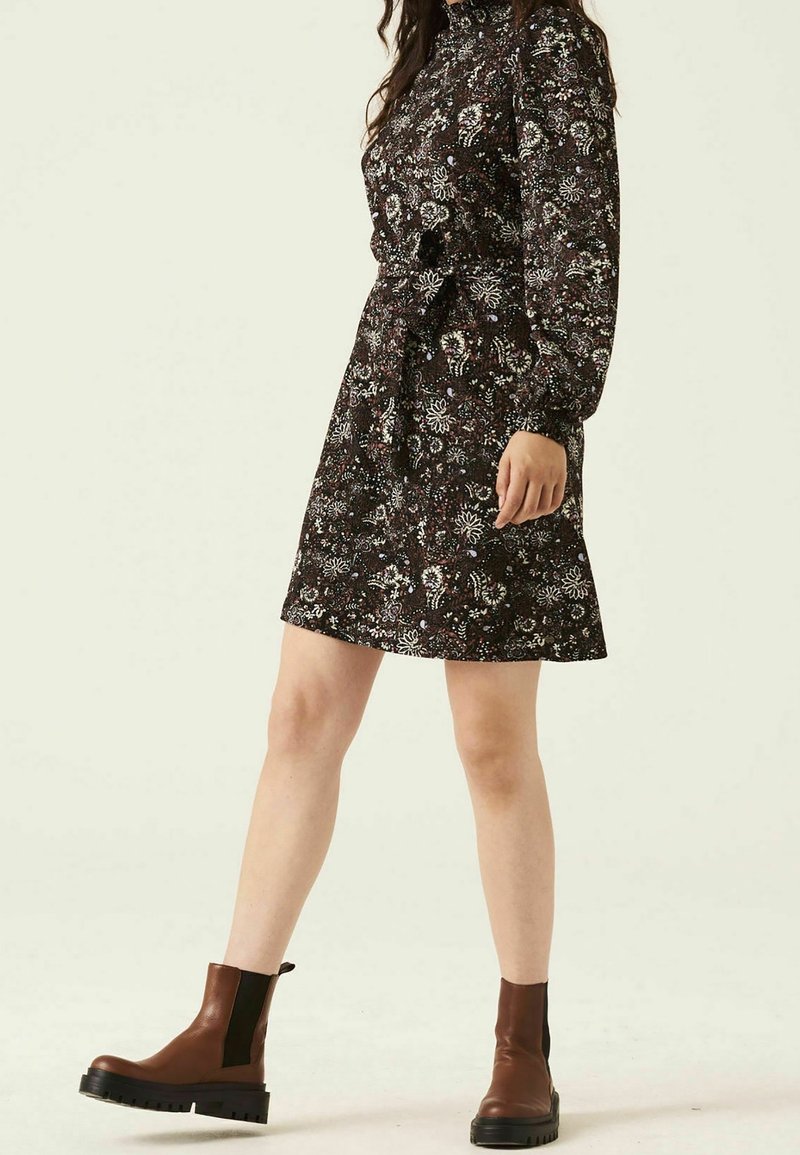 Schwarzes Kleid mit langen Ärmeln, floralem Muster, bindbarem Bund und ausgestellt Rock. Trägt dazu braune Stiefeletten mit dicker Sohle.