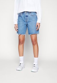Lichtblauwe denimshorts met rafelige zoom, hoge taille, gecombineerd met witte sneakers en sokken. Model draagt een wit overhemd met knopen.