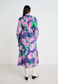 Långärmad blommig klänning med lila, rosa och gröna mönster. Materialet är strukturerat med en midja i bälte och en svävande silhuett.