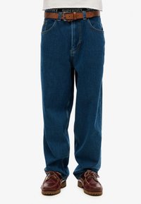 Vaqueros de mezclilla azul con un corte relajado, que incluyen un cinturón de cuero marrón. Los vaqueros tienen costuras prominentes y un diseño de cinco bolsillos. Unos zapatos marrones completan el look.