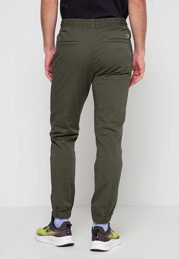 Pantalons de jogging vert olive avec taille élastique et deux poches arrière. Le matériau semble lisse avec une coupe décontractée, associés à des chaussures de sport.