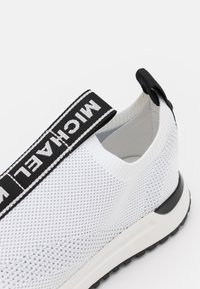 White knitted sneaker featuring a black logo strap, cushioned sole, and textured upper with a sleek design. Suitable for casual wear.