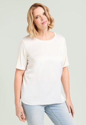 Donna con capelli biondi che indossa una maglietta bianca semplice a maniche corte e jeans azzurri, in piedi davanti a uno sfondo chiaro.