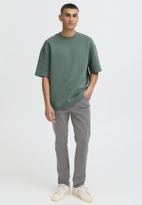 Zöld bővített fazonú rövid ujjú póló, világosszürke slim fit nadrággal és krémszínű, három csíkkal díszített tornacipővel párosítva.