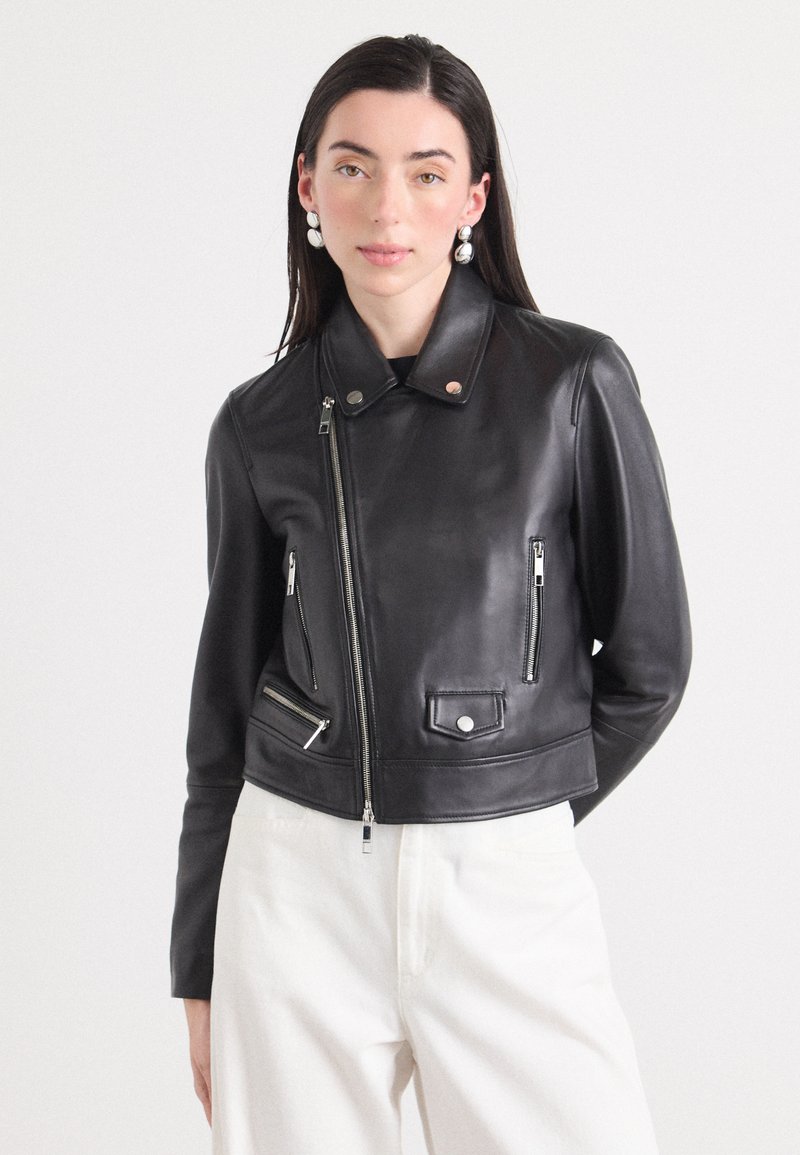 Model wears a black leather cropped jacket with silver zippers and pockets, paired with white pants. The background is plain and light.