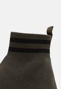 Zielona dziana skarpetka z wysokim kołnierzem, z czarnymi poziomymi paskami na górze. Tekstura jest gładka, a splot gęsty.