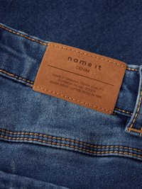 Syvänsinistä denimpukua, jossa on nahkainen merkki. Merkissä lukee "name it DENIM", ja siinä on tietoja laadusta ja perustamisajankohdasta, ommeltu kultaisella langalla.