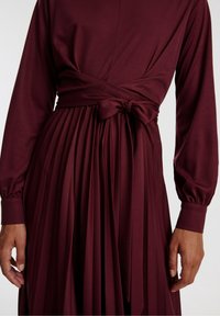 Burgundiru vestido con mangas largas y abullonadas y falda plisada. Presenta una cintura envuelta con un detalle atado, hecho de una tela suave.