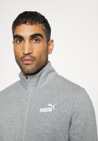 Giacca grigia con zip, caratterizzata da un colletto alto e dal logo Puma bianco sul petto. Il tessuto sembra morbido con una finitura testurizzata.