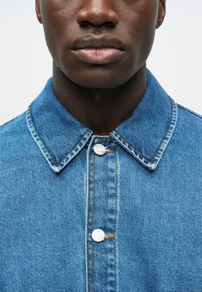 Zbliżenie na dolną część twarzy i szyję osoby noszącej niebieską kurtkę dżinsową zapiętą pod kołnierzem, z widoczym przeszyciem i srebrnymi guzikami.