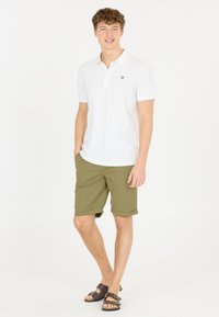 Witte poloshirt met korte mouwen en zwart logo, gecombineerd met olijfgroene shorts. Sandalen met bruine banden en gespen. Casual outfit.