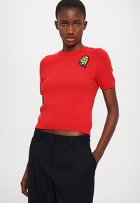 Maglione corto a maniche corte di colore rosso con orlo a coste e un dettaglio ricamato in nero e oro sul lato sinistro del petto. Si abbina a pantaloni scuri e sartoriali.