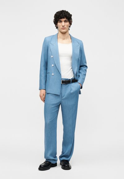 Jeune homme debout portant un costume croisé bleu clair, chemise blanche, ceinture noire et chaussures noires sur un fond uni.