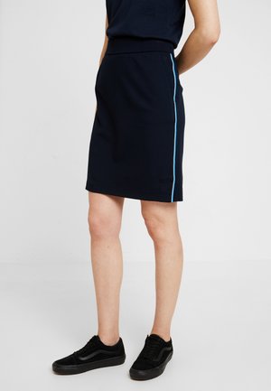 Personne portant une jupe bleu marine avec une fine bande latérale bleu clair, associée à des baskets noires, debout devant un fond clair uni.