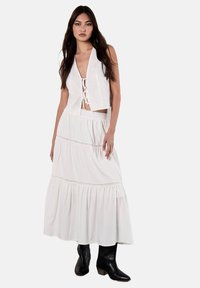 Top crocato bianco con dettagli ad occhiello e chiusura con lacci, abbinato a una lunga gonna maxi bianca a più strati. Stivali neri alla caviglia completano il look.
