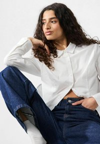 Camicia bianca con chiusura a bottoni, abbinata a pantaloni larghi in denim scuro. Tessuto morbido, design corto e vestibilità comoda.