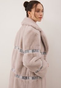 Futrzana kurtka w jasnym różu z szerokim kołnierzem i długimi rękawami. Posiada kontrastową, czarno-białą wstążkę z logo w okolicy talii i na rękawach.