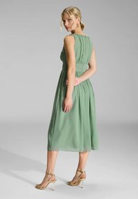 Robe verte, longueur genou, en tissu léger avec un dos plissé et une taille cintrée. Associée à des talons dorés à lanières. Arrière-plan uni.