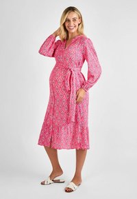 Robe rose à motifs avec des manches longues et un lien à la taille. Elle présente une jupe fluide avec un ourlet en volants, assortie à des sandales blanches.