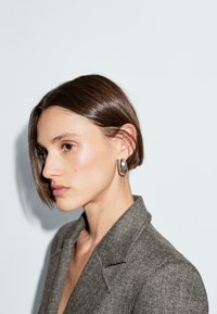 Szary wełniany żakiet z klapami noszony na gołej skórze. Modelka ma krótką, prostą fryzurę i nosi dużą, srebrną kolczyk.