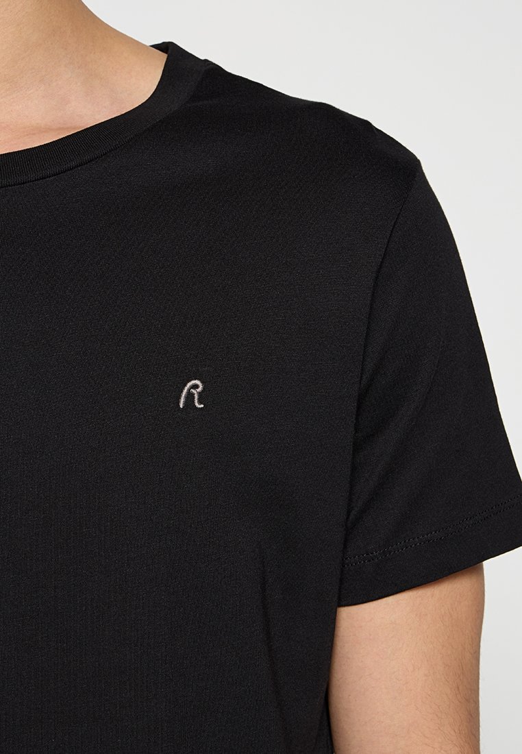 Schwarzes T-Shirt mit kurzen Ärmeln aus weichem Stoff, das ein dezentes graues gesticktes Logo auf der Brust und einen klassischen Rundhalsausschnitt präsentiert.