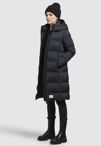 Dlhý tmavomodrý puffový kabát s kapucňou, vertikálnym prešívaním, bočnými vreckami a logom pri leme. Nosený cez čierne legíny a čižmy.