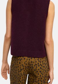 Bordowy bezrękawnik dzianinowy o teksturowym wykończeniu, w zestawieniu z brązowymi spodniami w czarne kropki i tylnymi kieszeniami.