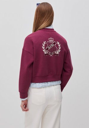 Kobieta z długimi włosami, ubrana w bordową bluzę z białym emblematem "Sport Club" i białe spodnie, widziana od tyłu.